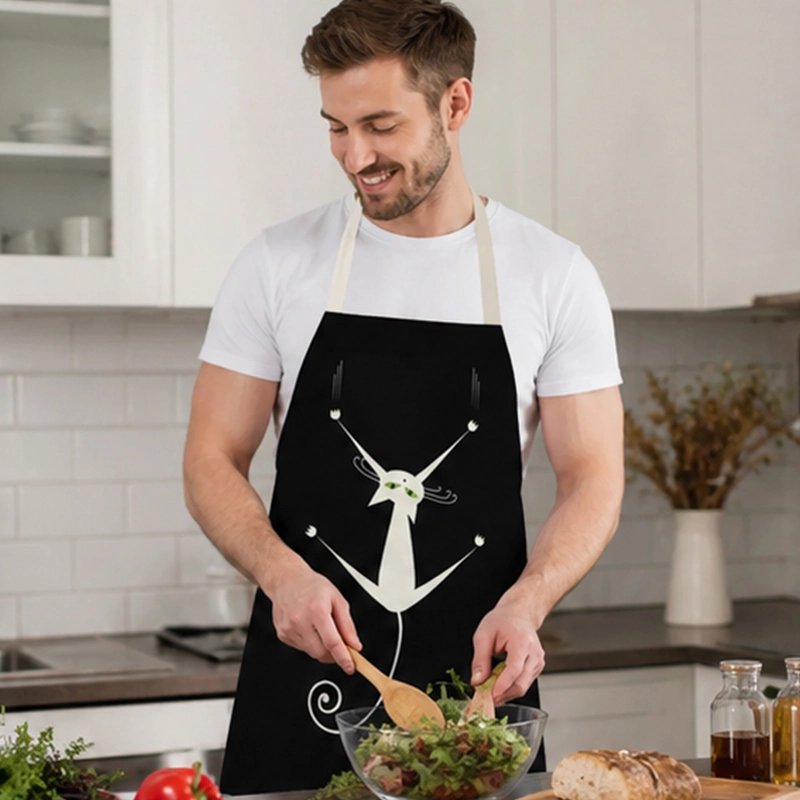 homme-dans-cuisine-prepare-salade-porte-tablier-noir-maintenu-par-des-lanieres-couleurs-claire-autour-du-cou-avec -motif-chat-blanc-au-centre